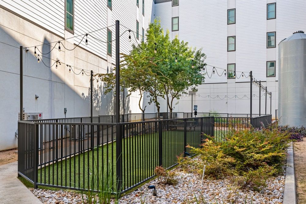 An urban outdoor dog park with a small, manicured grassy area and a tree, enclosed by a black metal fence. Overhead string lights drape between the building and the fence for ambient illumination. The building features a white facade with several windows, and a large metal silo-like structure is visible on the right side. The ground is covered with white pebbles and some shrubs in the foreground, suggesting a designed landscaping element at Marq on Burnet in Austin, TX.
