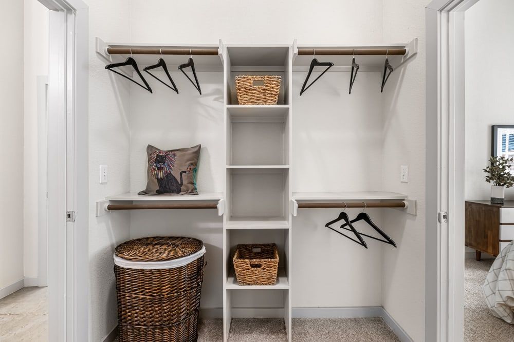 A neatly organized walk-in closet with white shelving and beige walls. Multiple shelves hold wicker baskets, and two clothing rods with black hangers are visible. On a lower shelf, there is a decorative pillow featuring a colorful bird design, and a matching wicker hamper sits on the carpeted floor to the left at Marq on Burnet in Austin, TX.