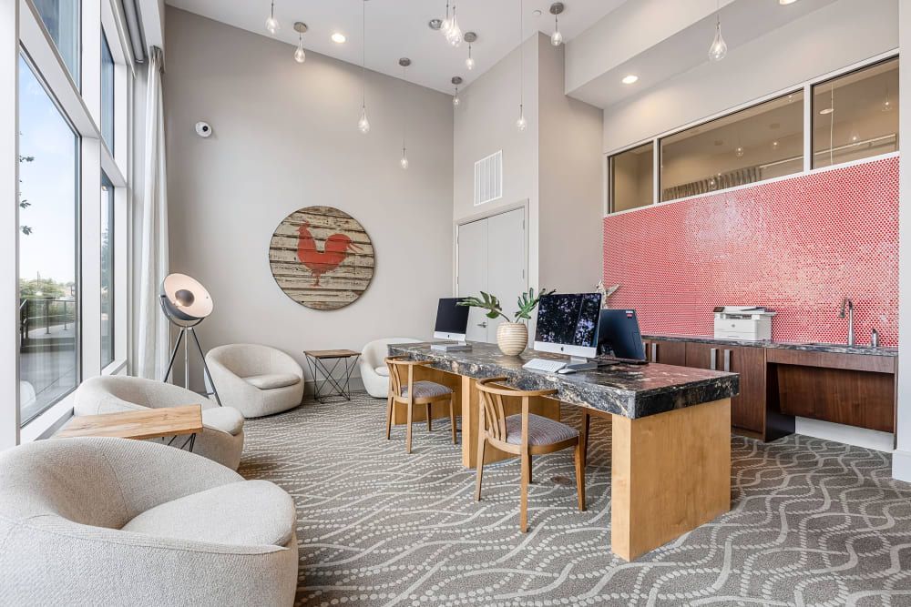 Modern office lounge area with gray carpet, two cream armchairs, a wood bench, and an artistic light fixture. There is a workspace with two computers on marble countertops against a red mosaic-tiled wall. A wooden circular art piece with a red heart is on the wall, and large windows provide natural light at Marq on Burnet in Austin, TX.