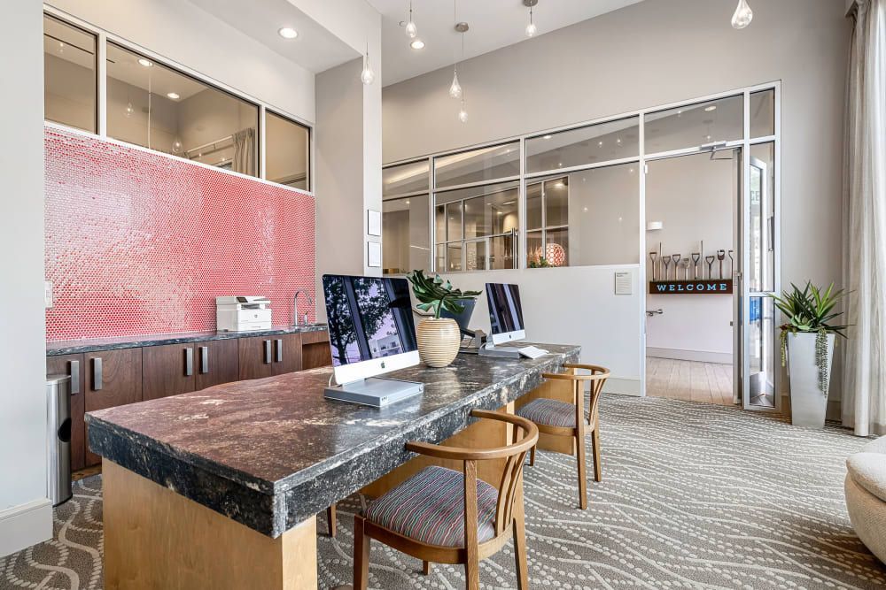 Modern and welcoming office with a desk featuring marble top, two iMac computers, and comfortable seating area. The interior design includes a tiled red backsplash, glass office doors with 'WELCOME' signage, pendant lighting, and decorative plants, creating a professional yet inviting atmosphere at Marq on Burnet in Austin, TX.