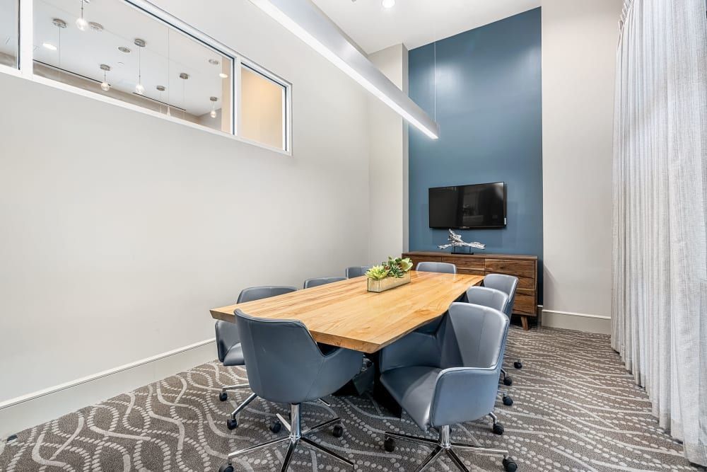 Modern corporate meeting room interior with a long wooden table, surrounded by blue upholstered chairs, a blue accent wall with a mounted TV, and a decorative window allowing natural light to illuminate the space at Marq on Burnet in Austin, TX.