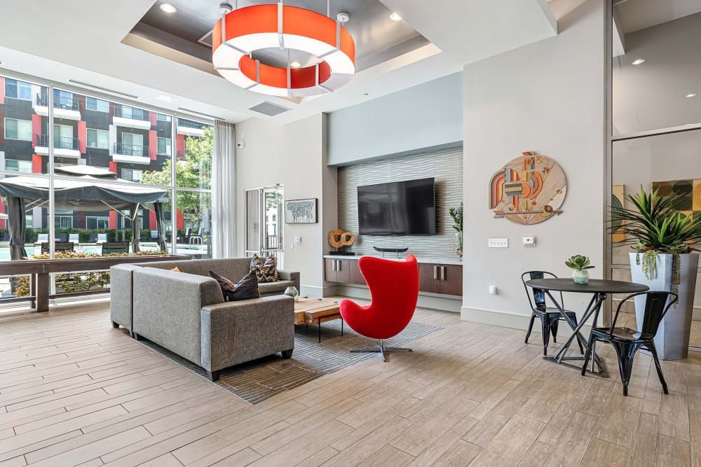 Modern apartment lobby with stylish decor, featuring a gray sofa, red accent chair, black dining table with chairs, and a mounted television. Large windows provide a view of the exterior building facade. Decorative art and plants add a cozy ambiance to the space. The room is well-lit with a large contemporary red and white hanging light fixture at Marq on Burnet in Austin, TX.