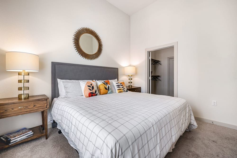 A cozy modern bedroom featuring a queen-size bed with a gray upholstered headboard and a white and gray geometric-patterned duvet. The bed is flanked by two wooden nightstands, each with a stylish table lamp with a gold and white cylindrical base. Above one nightstand hangs a round sunburst mirror. To the right, there is an open closet with built-in shelves partially visible. Decorative pillows add a pop of color, and the room is carpeted in a neutral tone, complementing the overall clean and inviting aesthetic of the space at Marq on Burnet in Austin, TX.