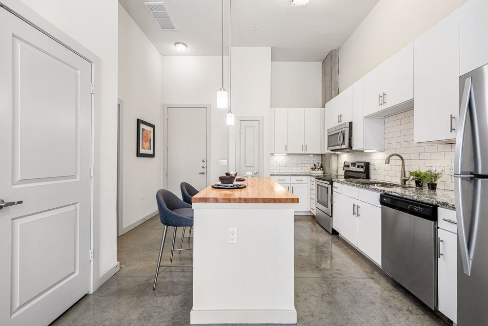 Modern kitchen interior with white cabinetry, stainless steel appliances, subway tile backsplash, and a central island with bar stools. The room features high ceilings, a concrete column, pendant lighting, and minimalistic decor with a framed artwork on the wall. The palette is neutral with white, grey, and touches of wood at Marq on Burnet in Austin, TX.