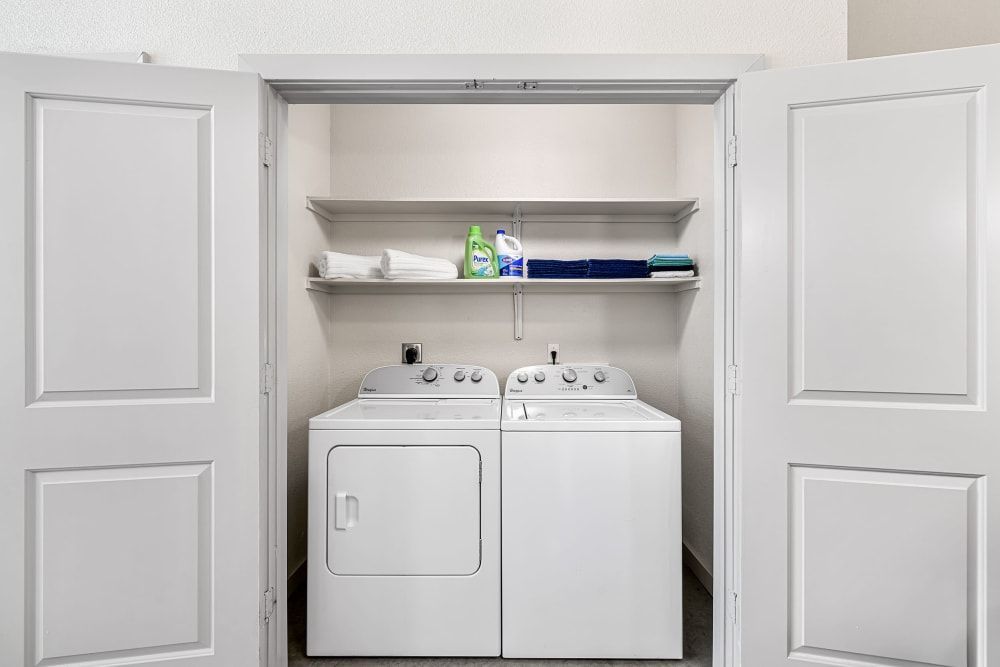 A clean and organized laundry closet with open white paneled doors revealing a white top-loading washing machine and a matching front-loading dryer. Above the appliances are shelves holding a small stack of folded white towels, a blue towel, and a bottle of liquid laundry detergent. The walls and appliances are in various shades of white, suggesting a bright and neutral space at Marq on Burnet in Austin, TX.