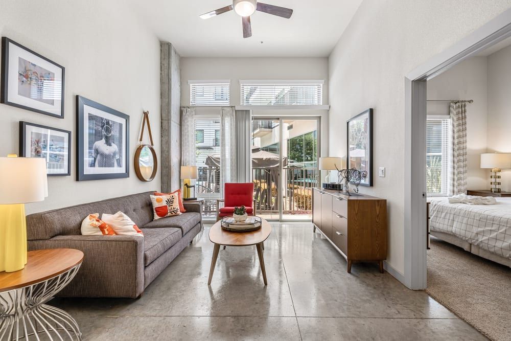 Modern living room in an apartment with a gray upholstered sofa adorned with orange accent pillows, wood-topped round coffee table, and a single red armchair. The room features a high ceiling with a ceiling fan, concrete support column, and framed artwork on the walls. To the right is a glimpse into a bedroom with a made bed and a sliding door leading to a balcony with outdoor furniture. Natural light fills the space, creating a bright and airy ambiance at Marq on Burnet in Austin, TX.