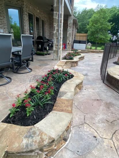 A patio with a stone planter filled with flowers and chairs.