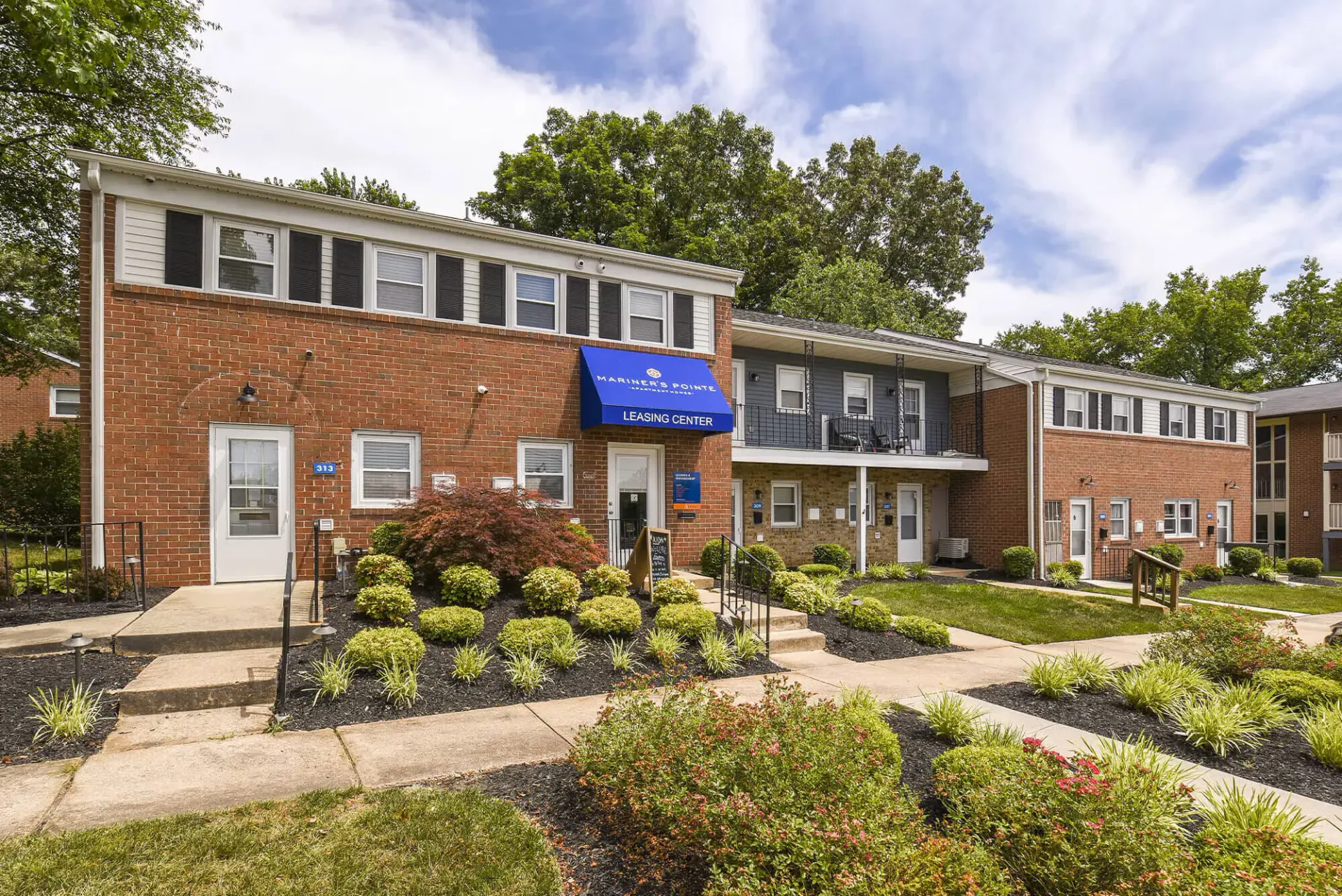 Exterior view of a brick apartment community with a blue leasing center sign and landscaped walkways.