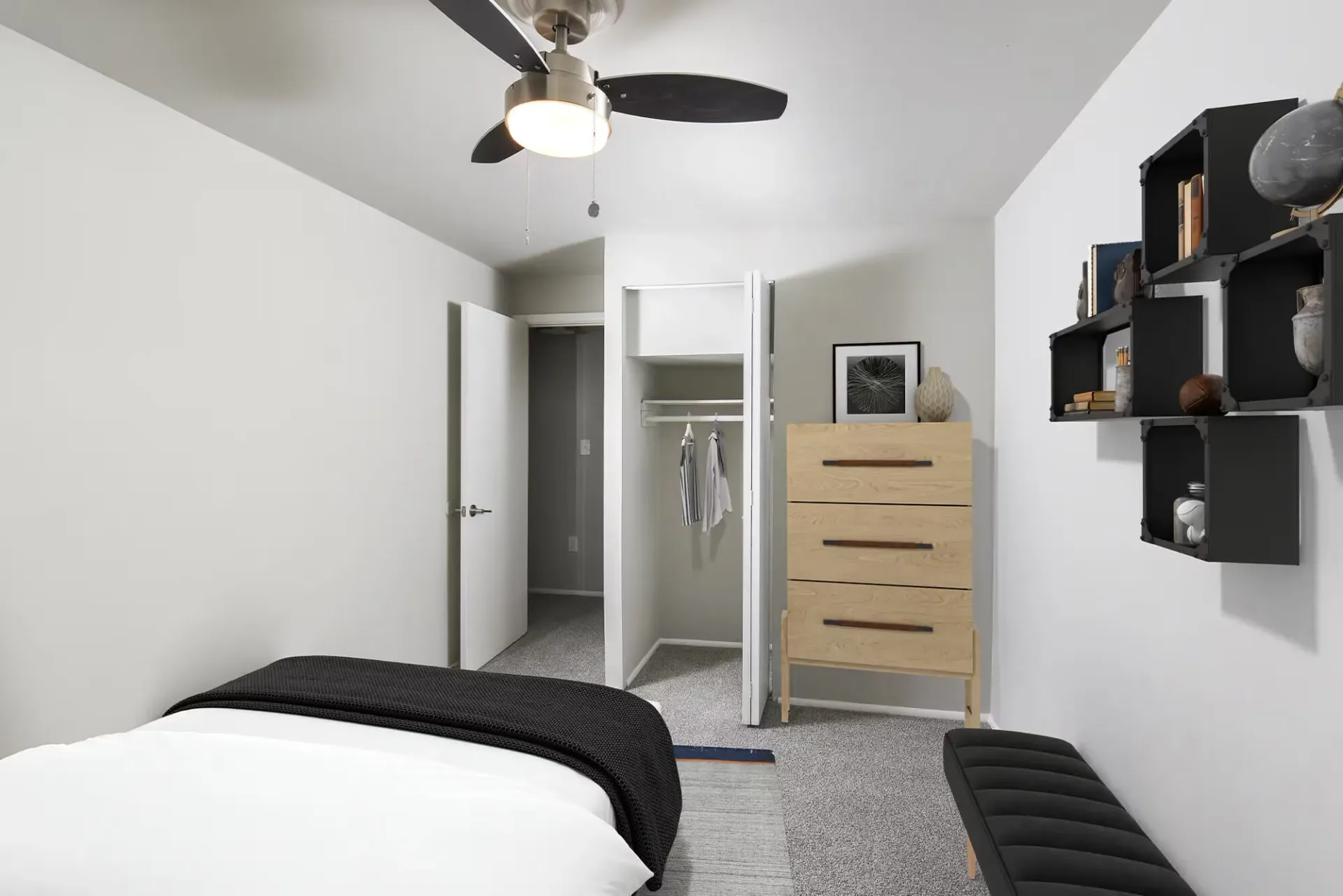 Bedroom in an apartment with an open closet, a light wood dresser, wall-mounted shelves, a bench, and a ceiling fan.