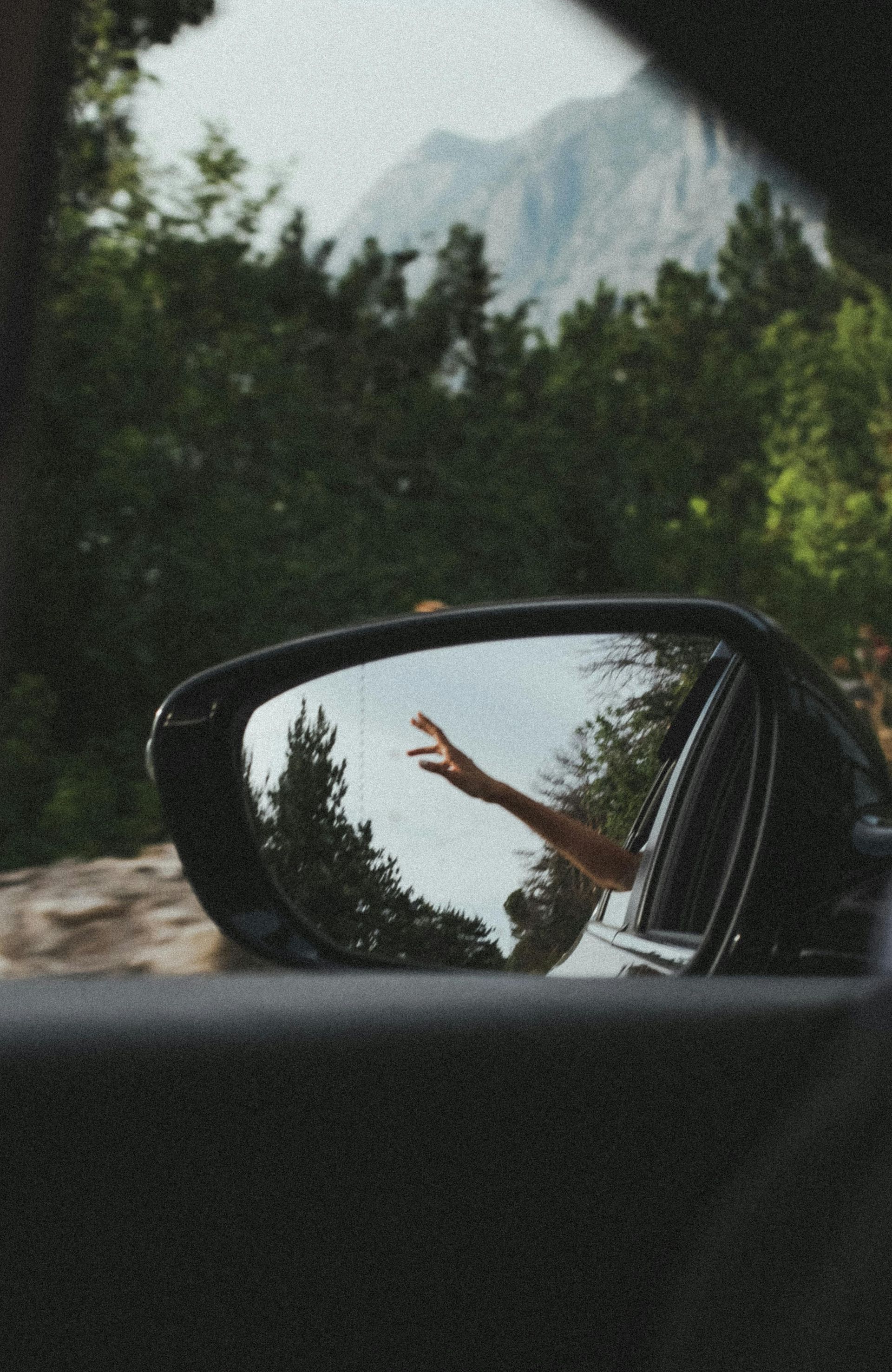 Car side mirror with a hand reaching out, reflecting trees, mountains, and sky.