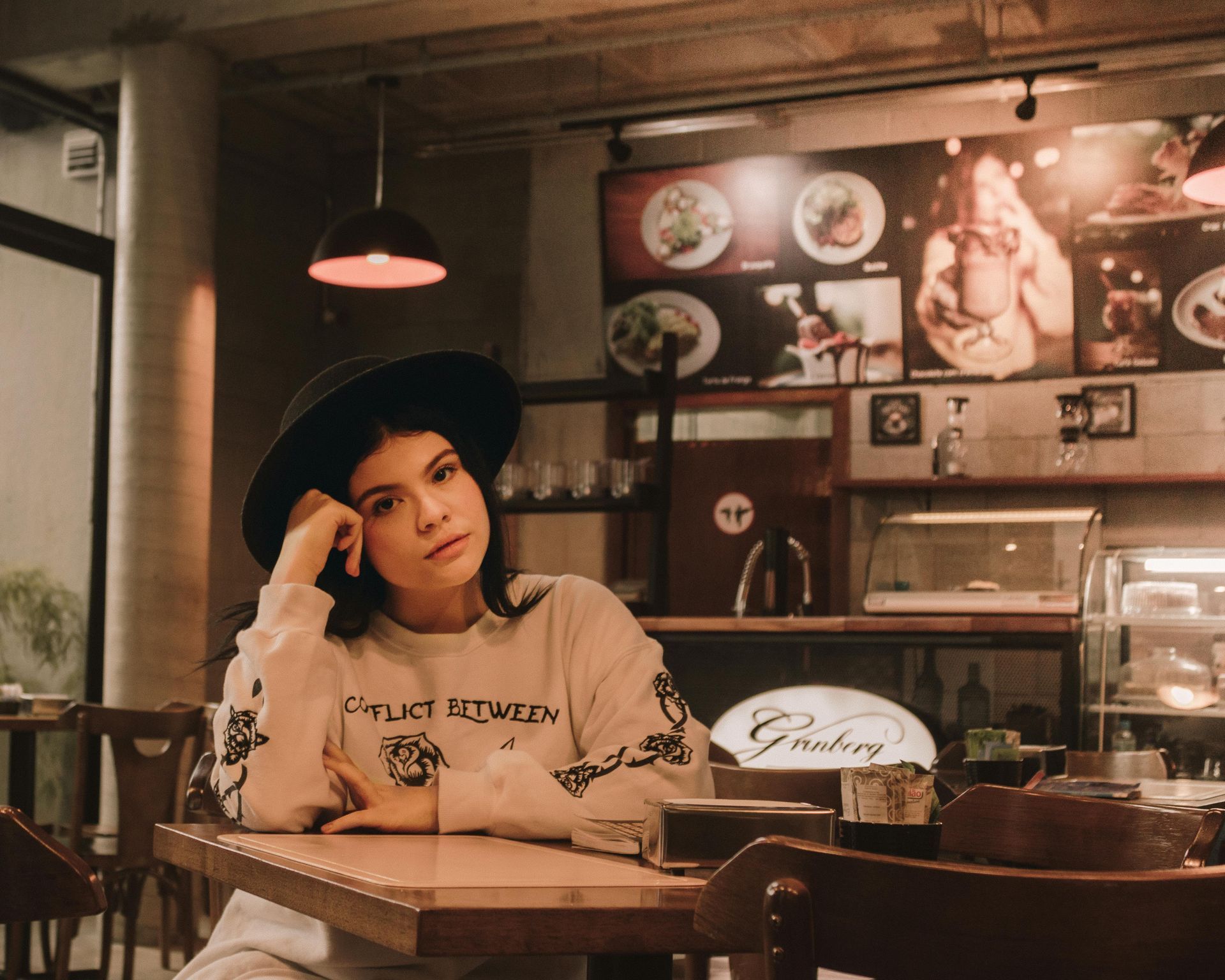 Woman in black hat and sweater at a cafe, resting head on hand. Wooden table, menu board in background.