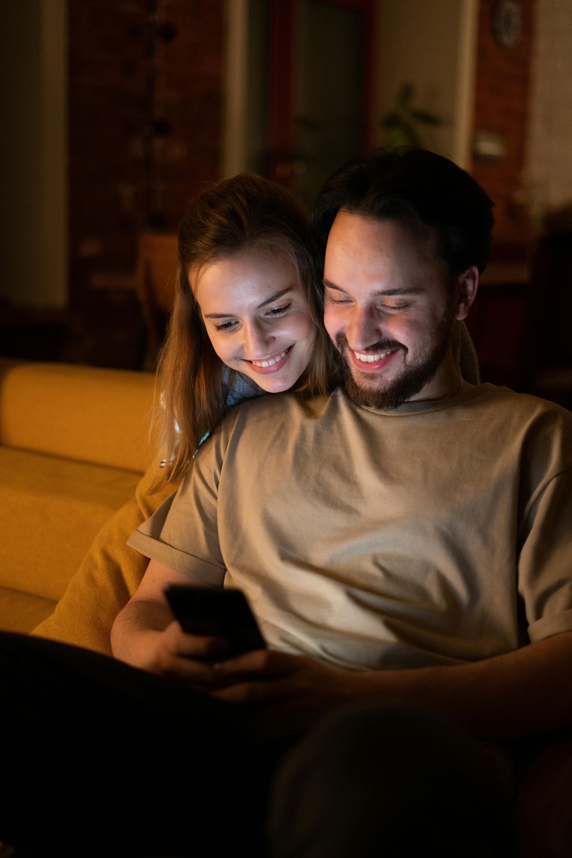 Couple smiling, looking at phone together, sitting on couch in a dimly lit room.