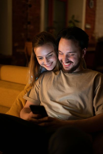 Couple smiling, looking at phone together, sitting on couch in a dimly lit room.