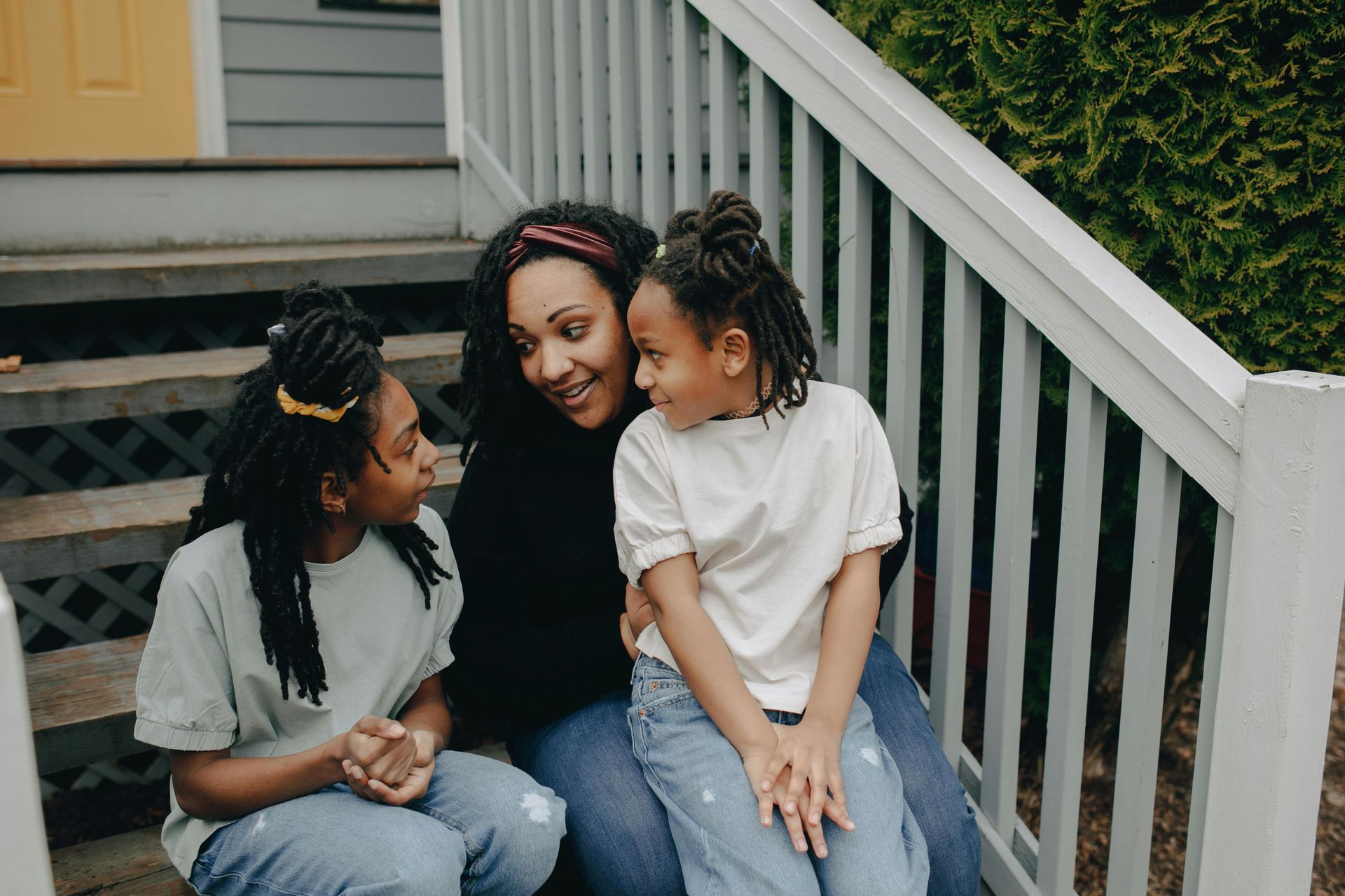 Woman sitting on steps, smiling at two children. They sit close together. White stairs.