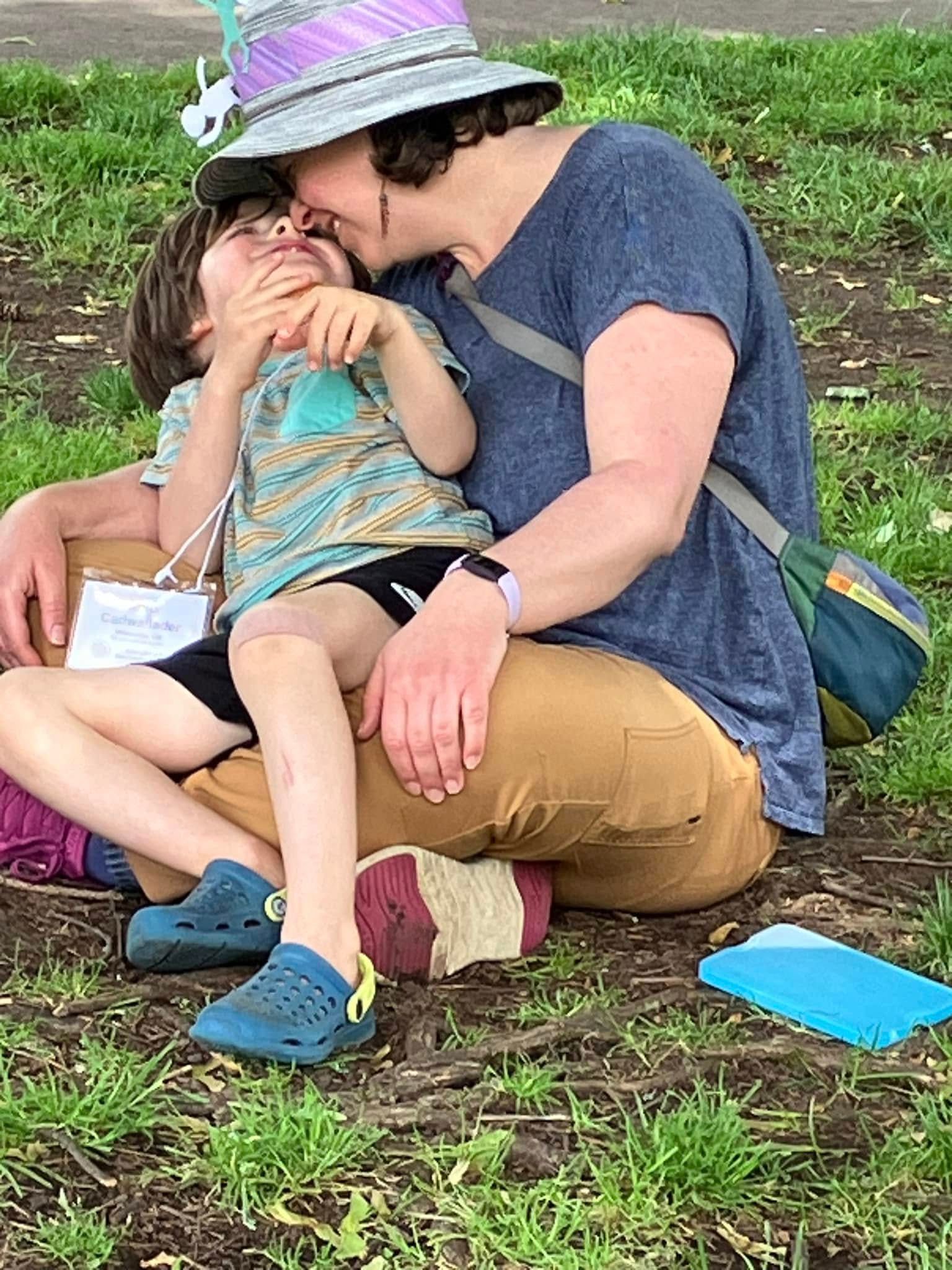 Mother wearing glasses smiling with eyes closed while holding a child wrapped in a blue blanket, both resting closely together on a striped chair in a sandy outdoor setting.