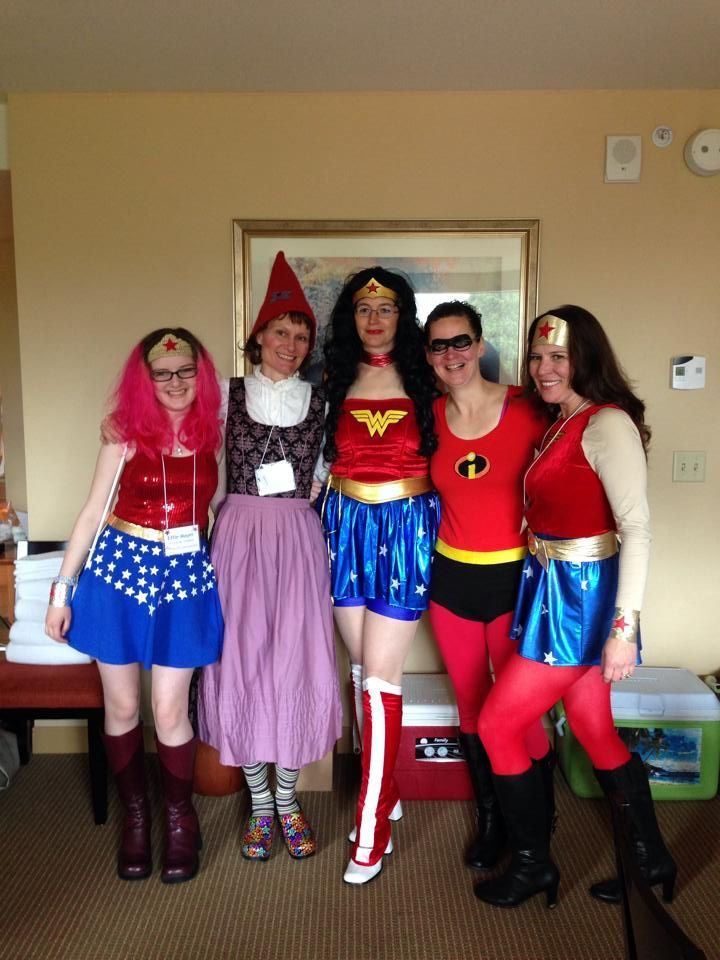 Five people in colorful costumes, including Wonder Woman and a gnome, smiling in a cozy room.