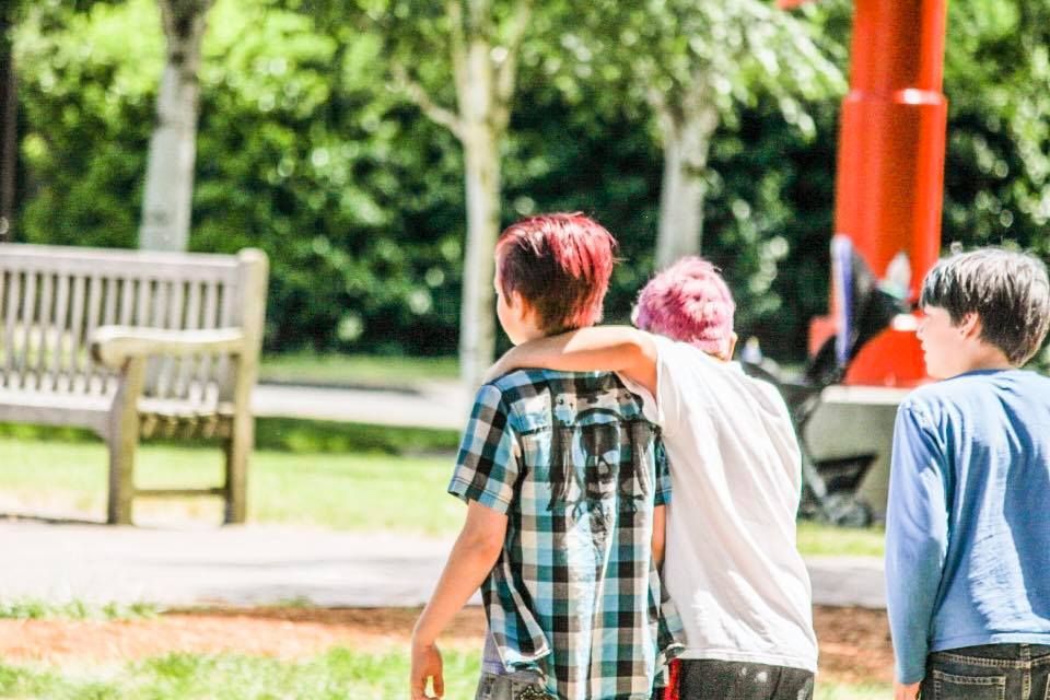 Group of thirteen unschoolers standing together on a grassy area, smiling and posing for a photo in a park-like setting with trees and buildings in the background.