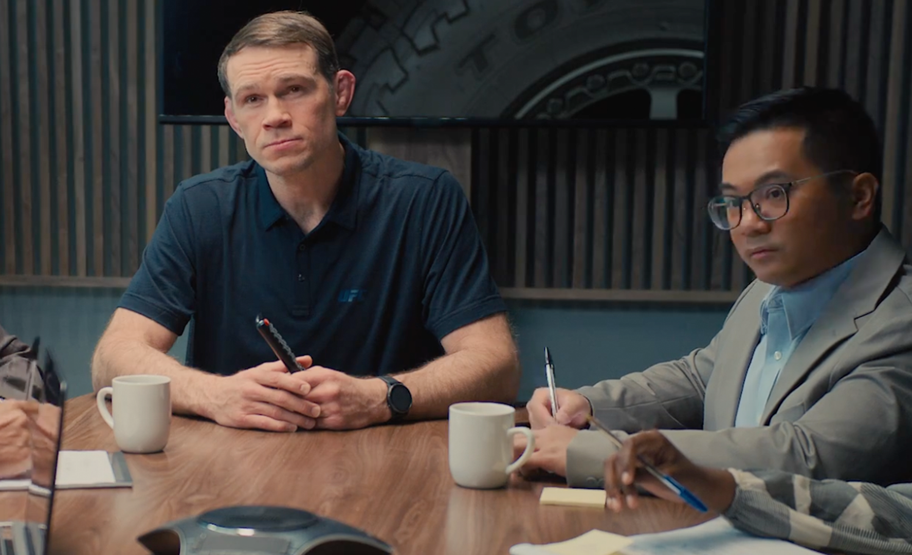 Two men at a conference table, listening and taking notes. The man on the left has a serious expression.