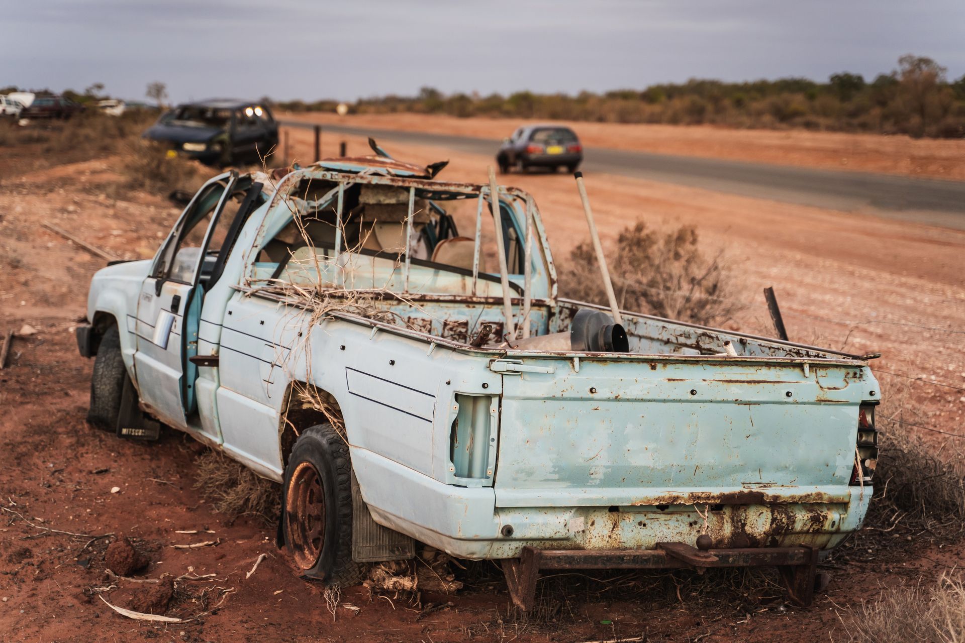 A dilapidated, white pickup truck on the side of a rural road, with other vehicles visible in the distance.