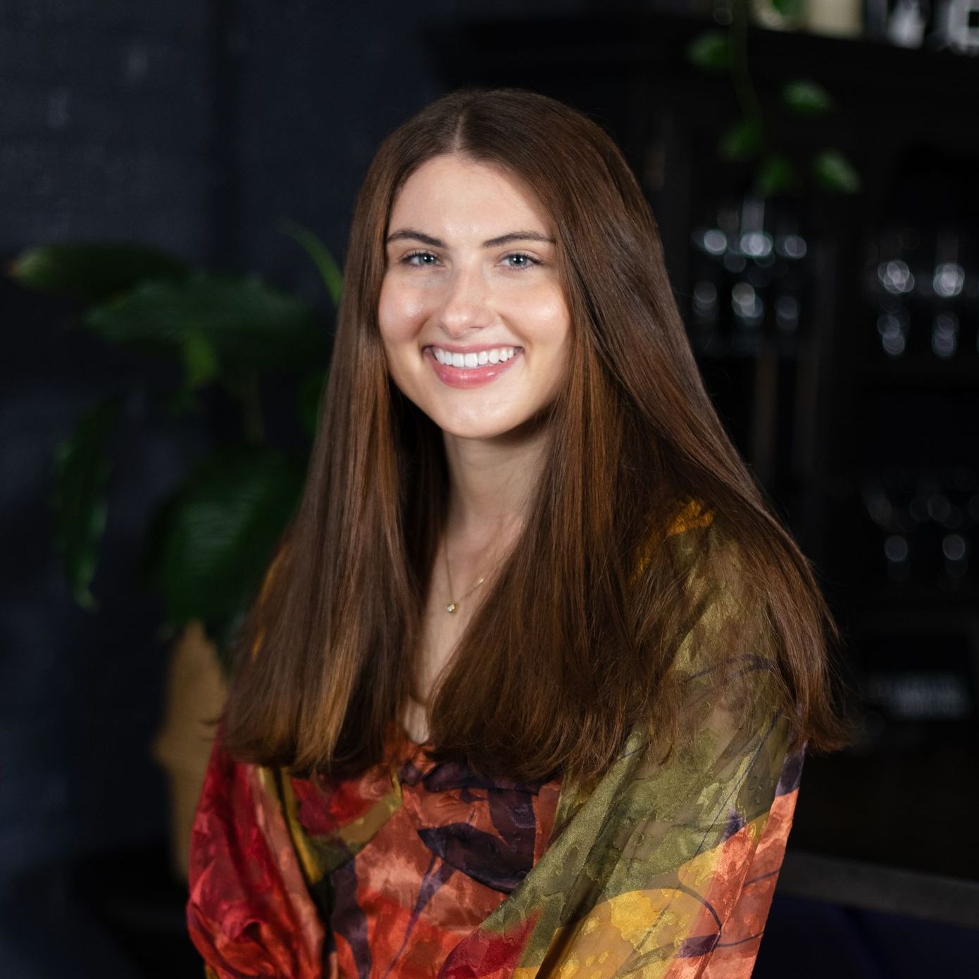 A woman in a red shirt and white pants is sitting on a stool and smiling.