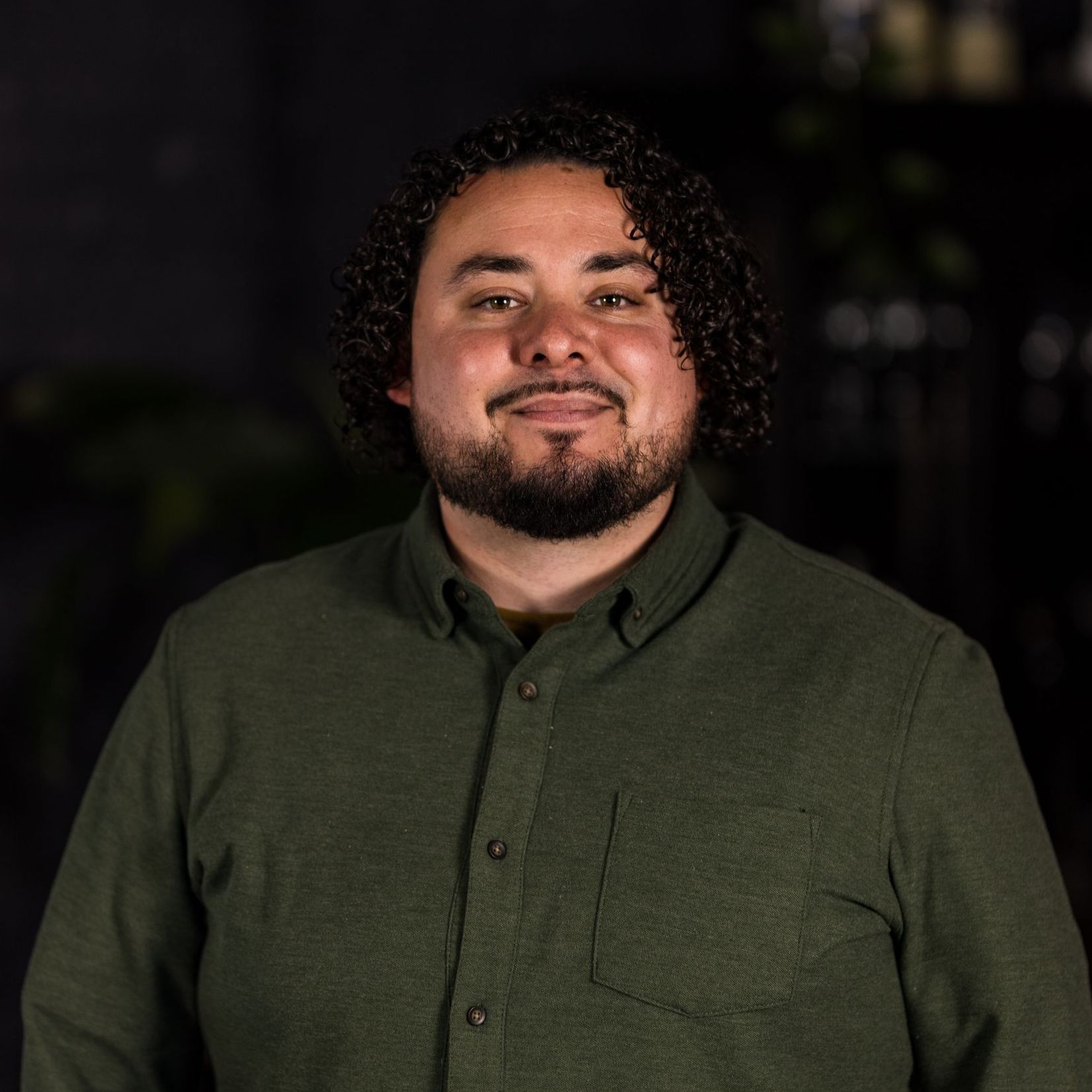 A man with curly hair and a beard is wearing a green shirt and smiling for the camera.