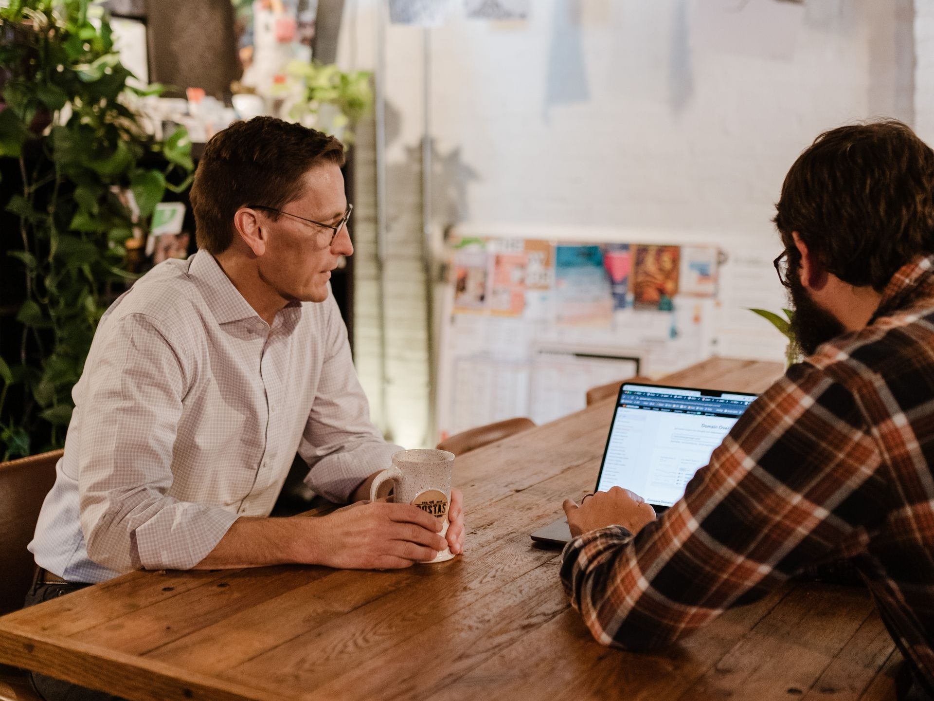 Two men are sitting at a table with a laptop and talking to each other.