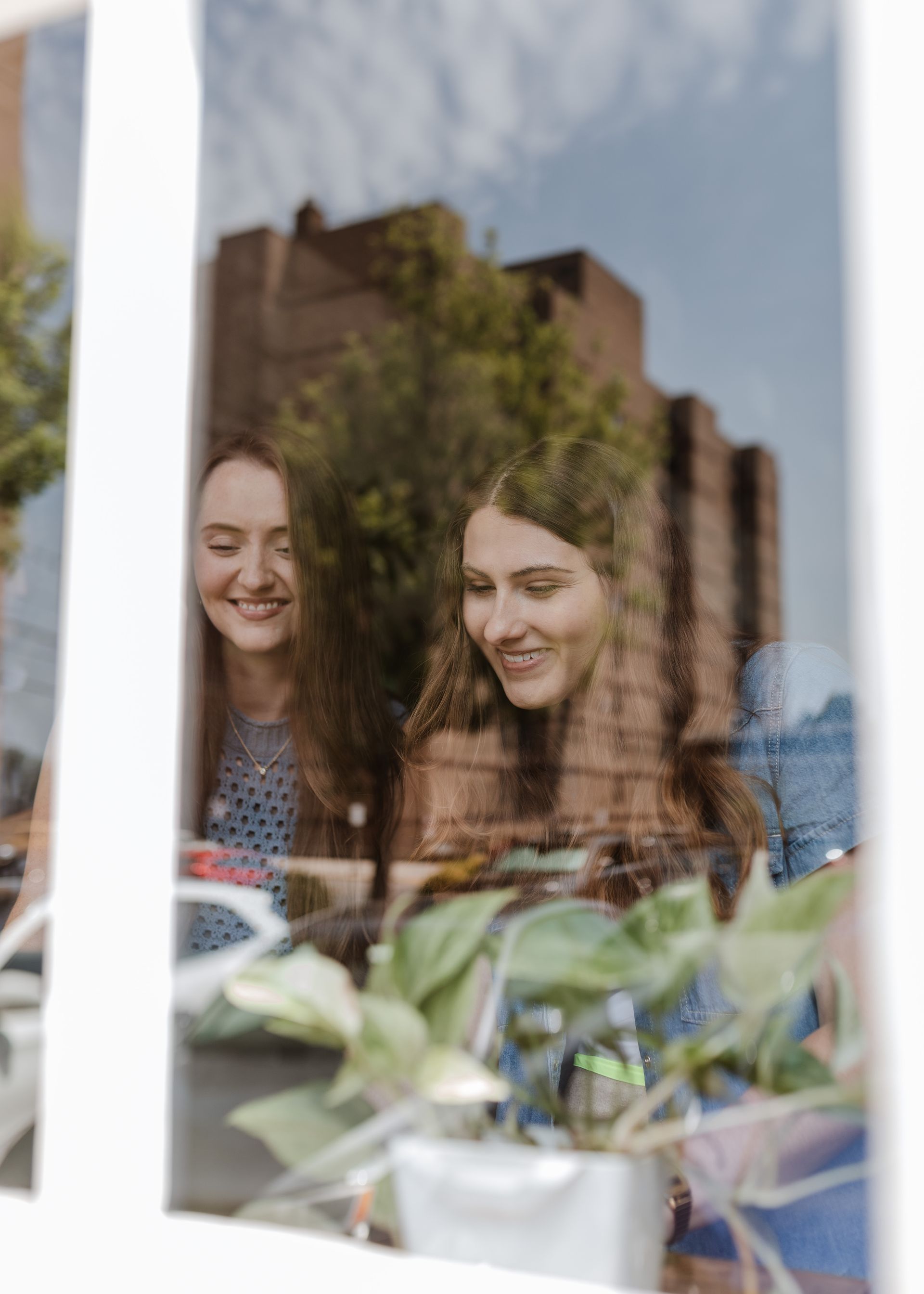 Two women are looking out of a window at a plant.