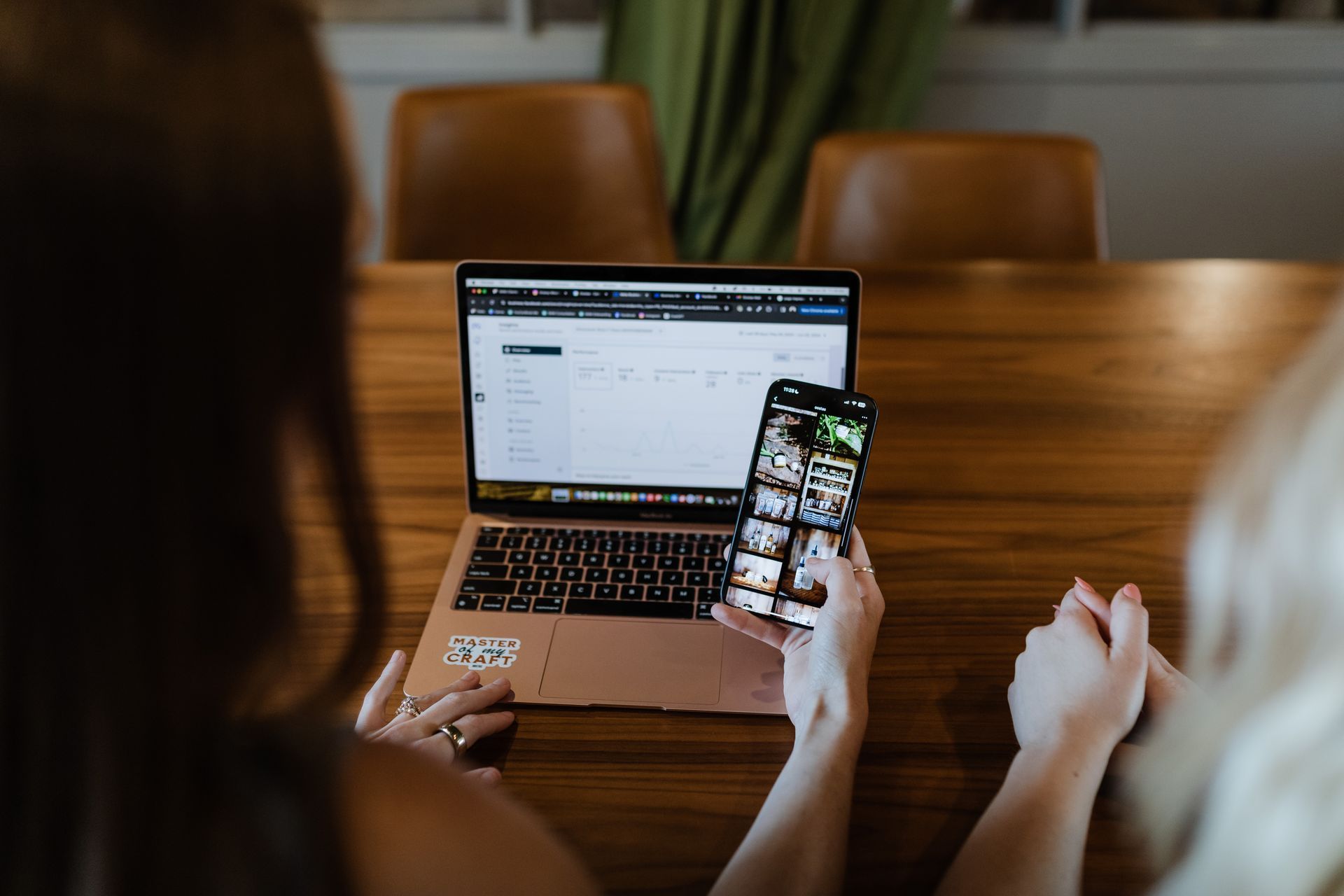 Two women are sitting at a table using a laptop and a cell phone.