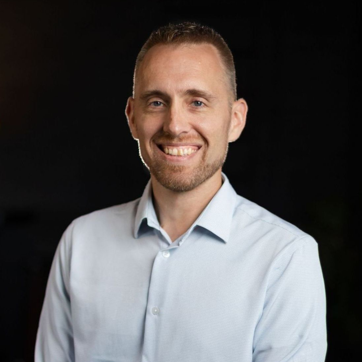 A man in a light blue shirt is smiling for the camera.