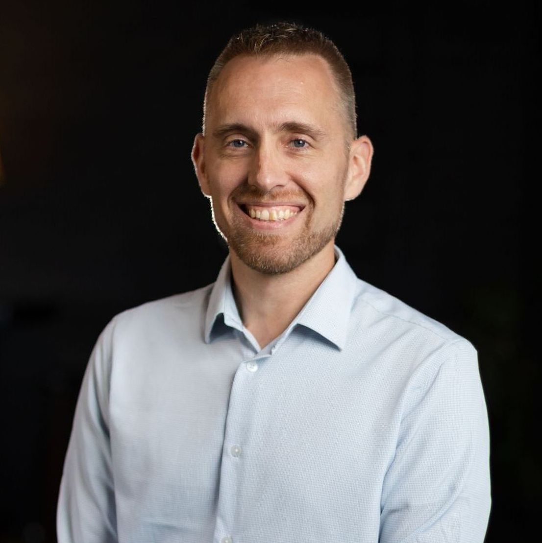 A man in a light blue shirt is smiling for the camera.