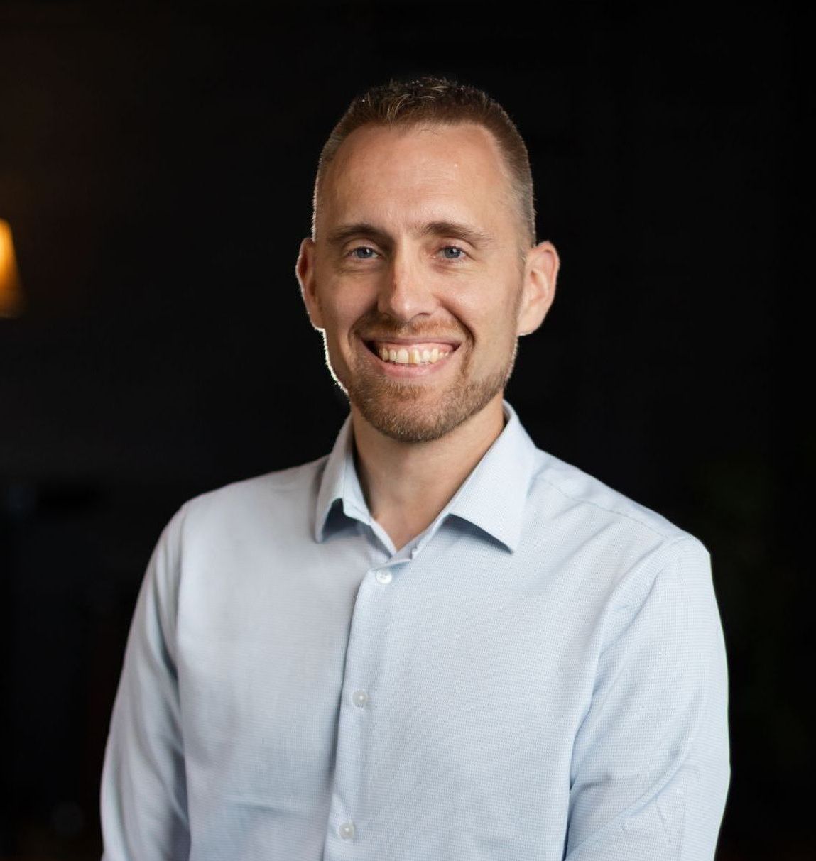 A man in a light blue shirt is smiling for the camera.