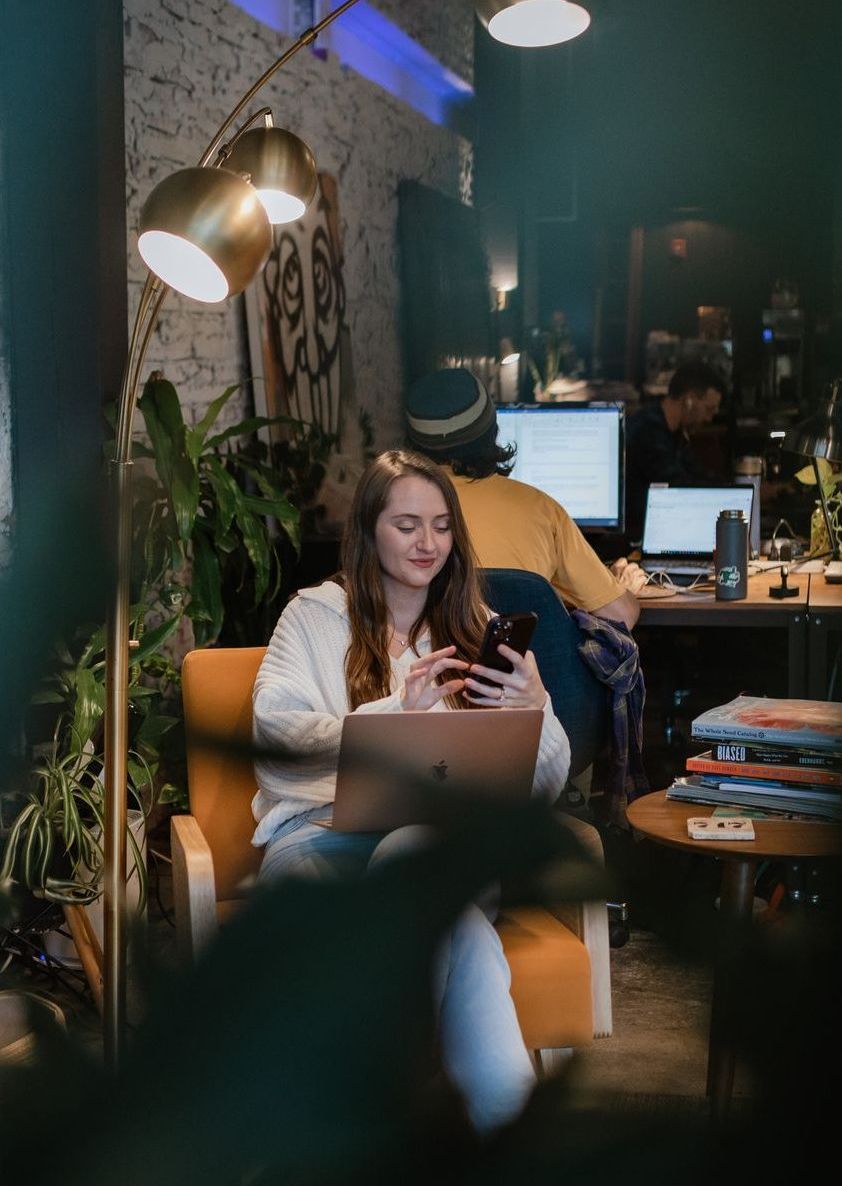 A woman is sitting in a chair using a laptop and a cell phone.