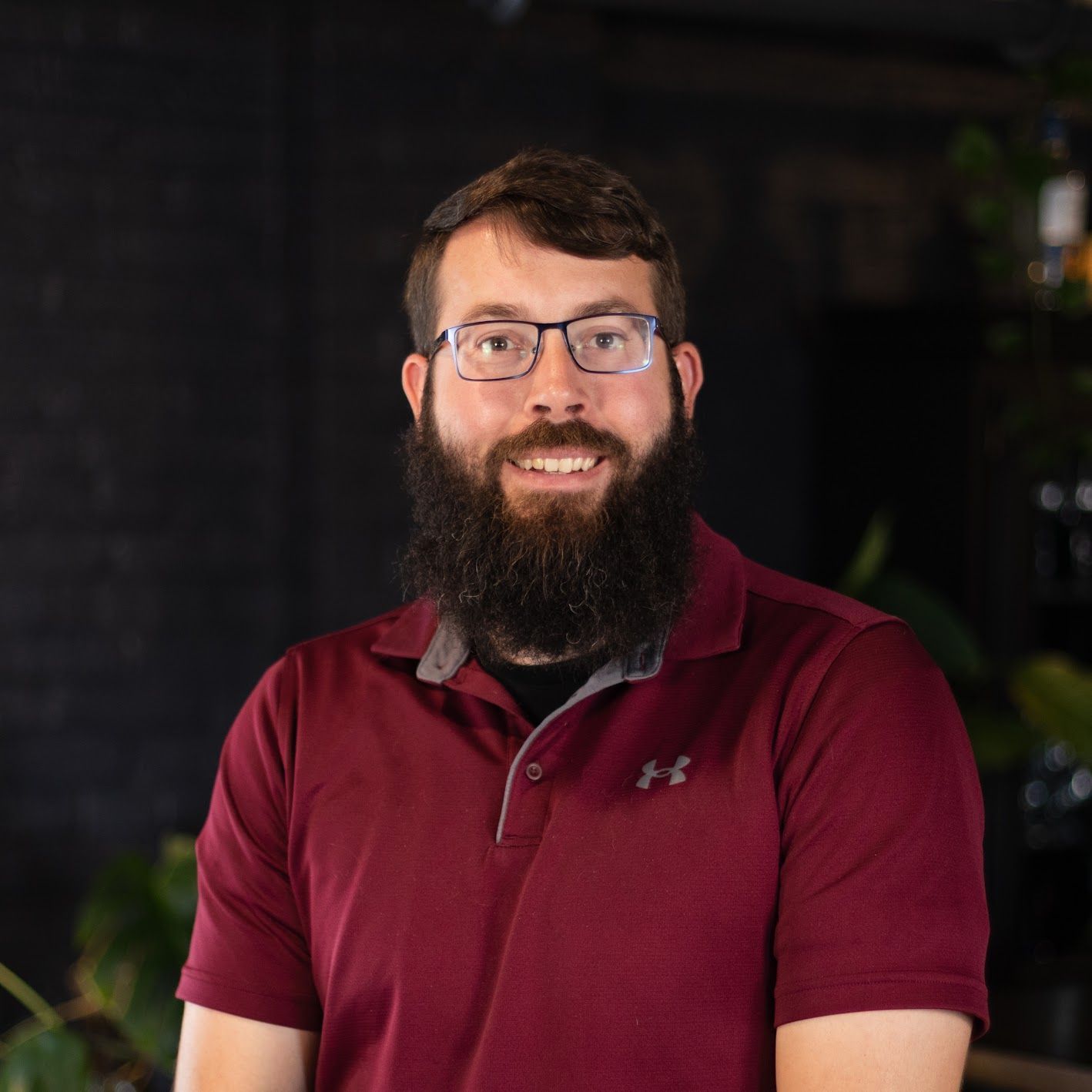 A man wearing glasses and a plaid shirt is sitting on a stool.