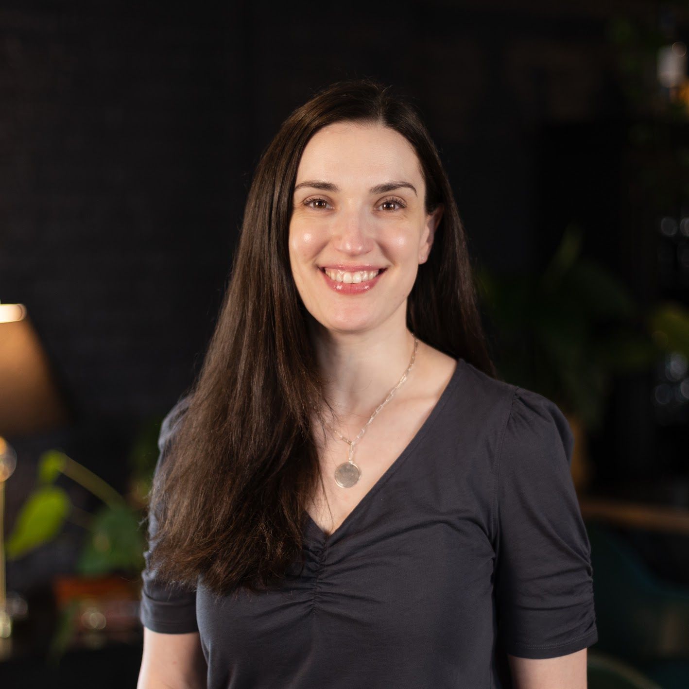 A woman in a blue shirt is sitting on a stool and smiling.