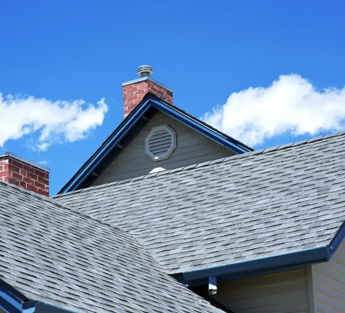 Tan house with multiple gabled roofs, dark gray shingles, and brick chimneys against a bright blue sky with light clouds.