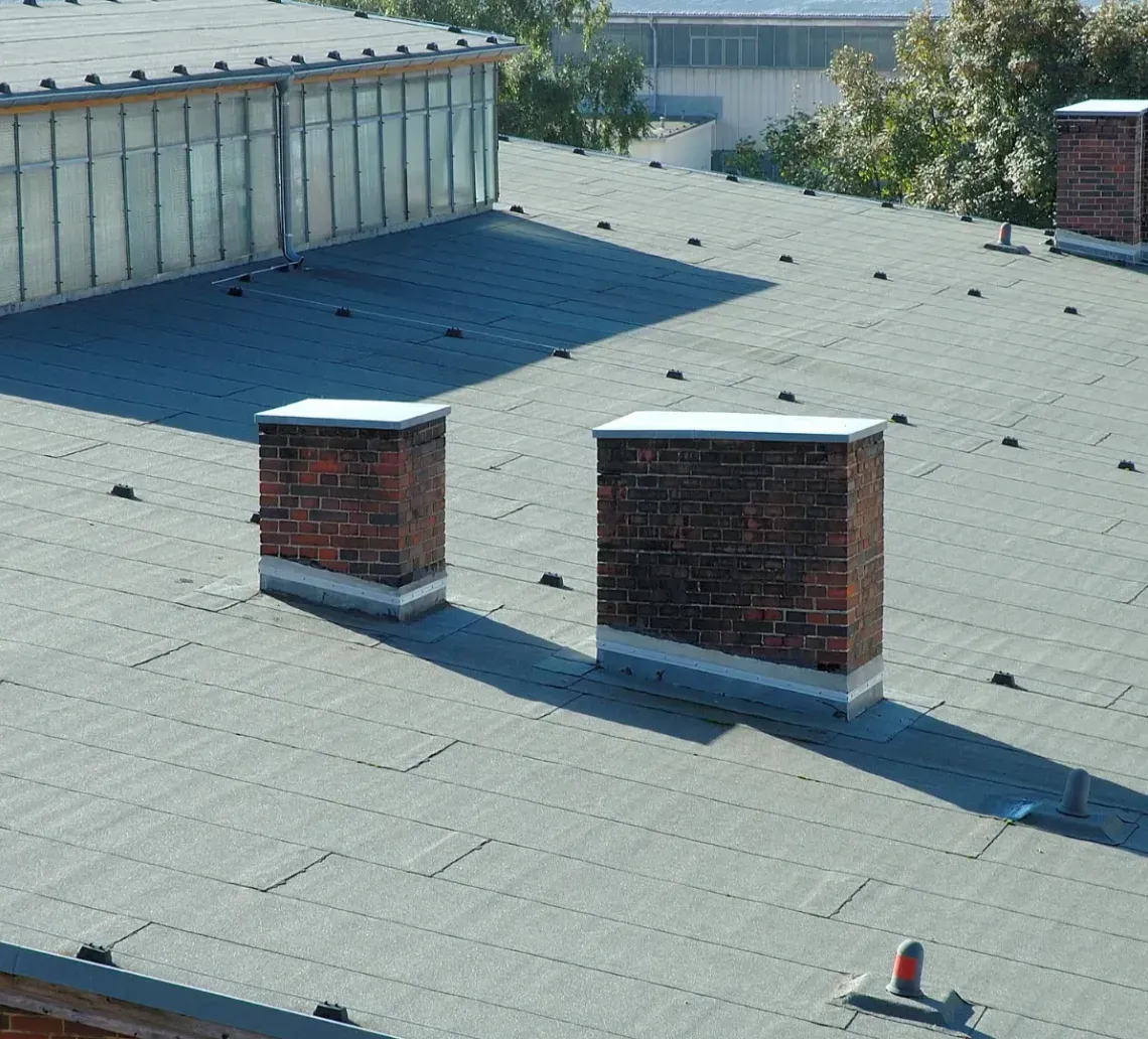 A gray, sloped roof featuring two red brick chimneys, a glass skylight structure, and several small ventilation pipes.
