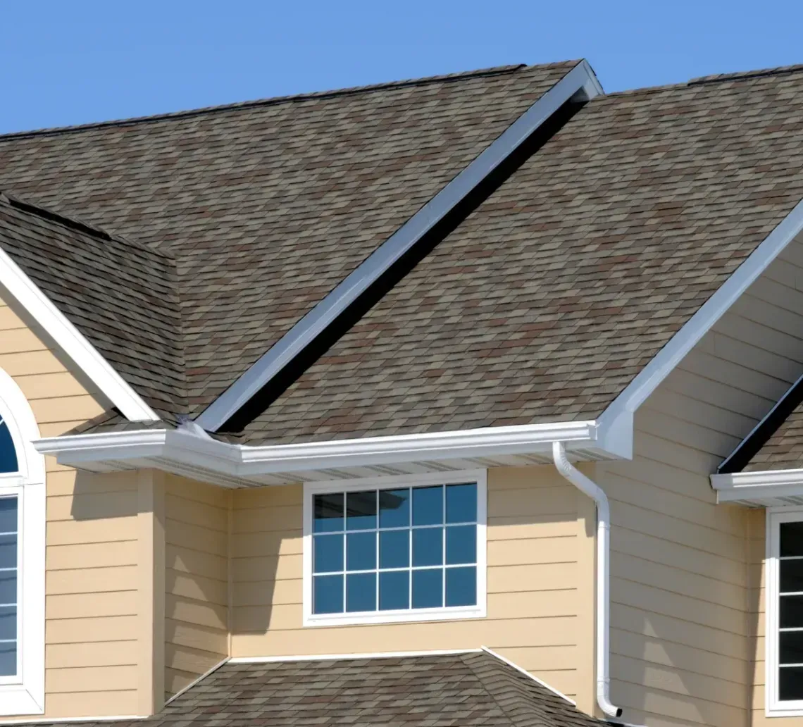The upper exterior of a two-story tan house with multi-gabled brown shingled roofs and white-trimmed windows.