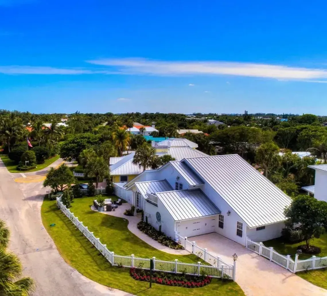 Aerial view of a coastal suburban neighborhood featuring a large white house with a metal roof and lush greenery.