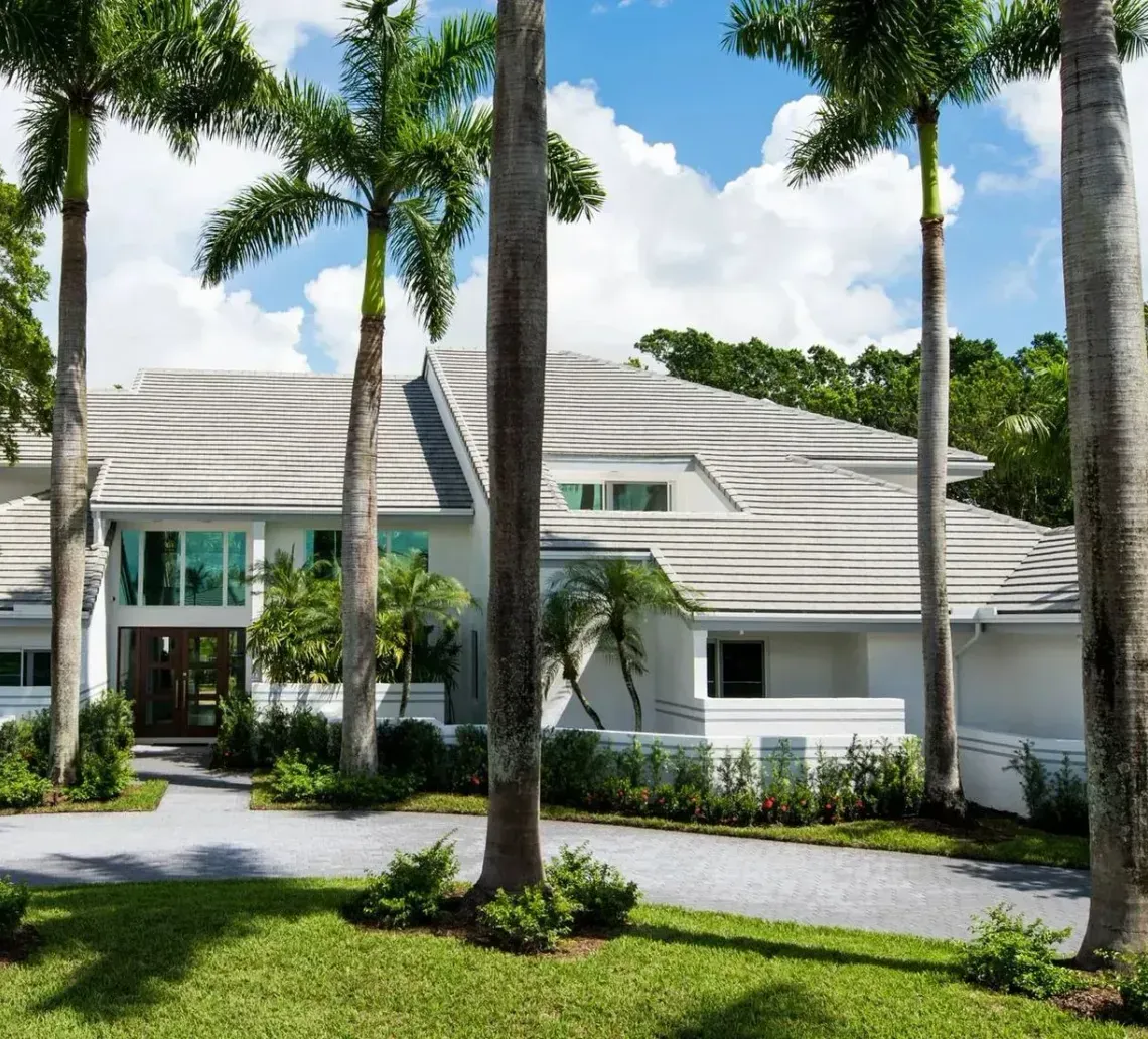 A large, multi-story white house with a layered roof, nestled among tall palm trees and lush greenery on a sunny day.