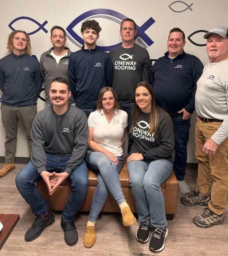 A group of ten people stand and sit in front of an office wall featuring a Oneway Roofing logo.