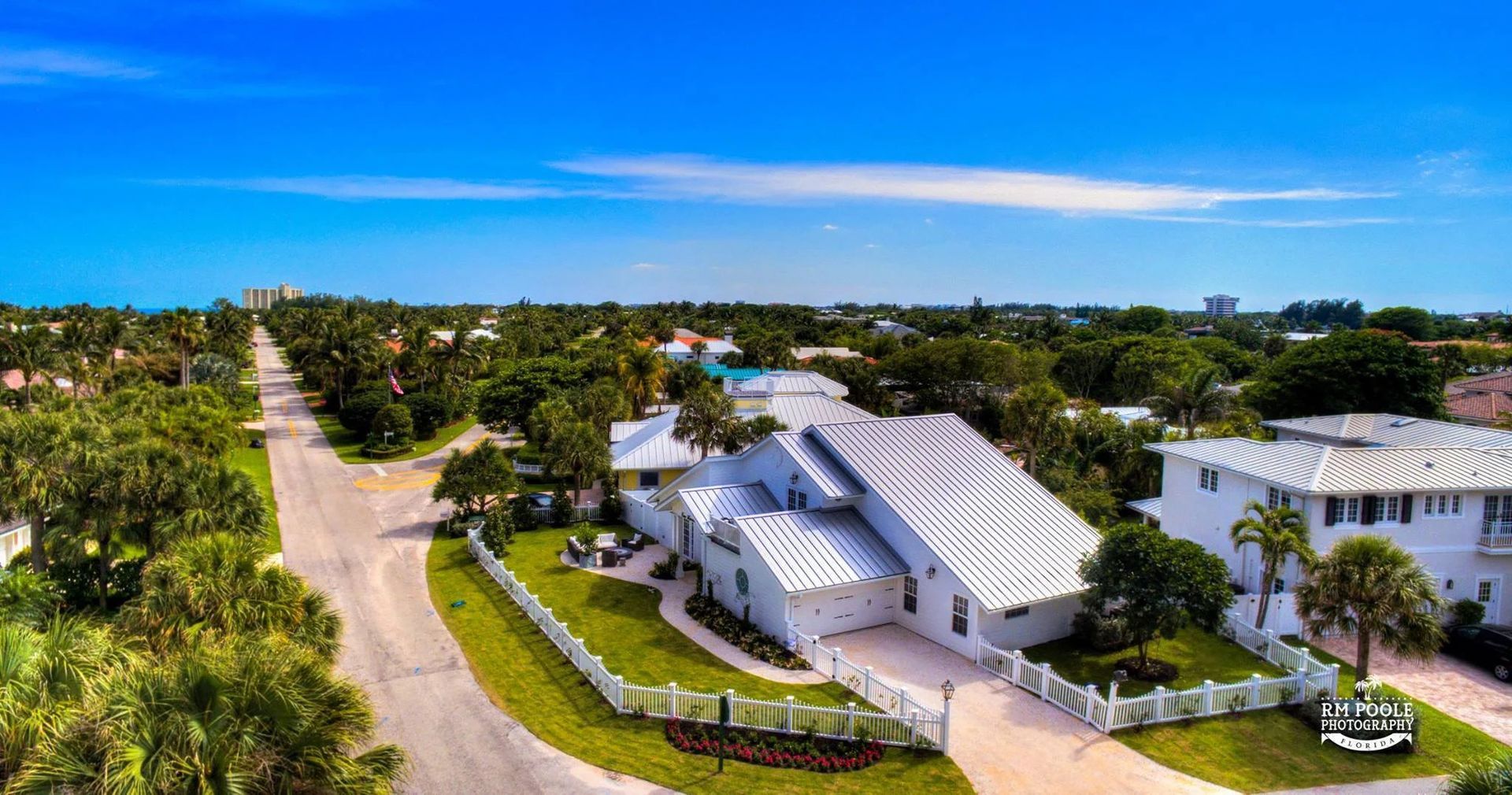 An aerial view of a white coastal home with a metal roof, surrounded by green trees and palm trees, along a paved road.
