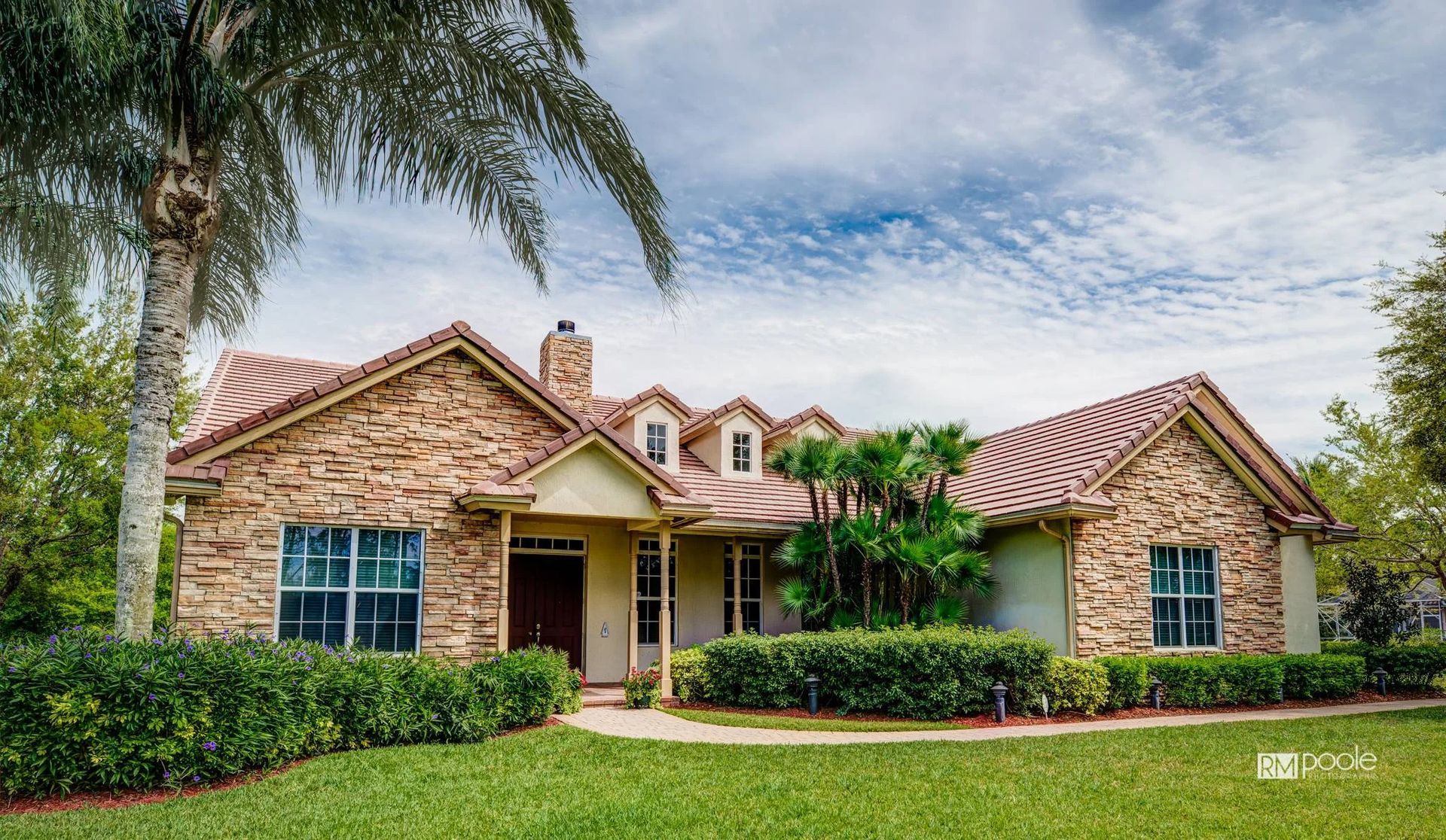 A one-story tan stone house with a multi-gabled, wavy red tile roof, surrounded by landscaping and a palm tree.