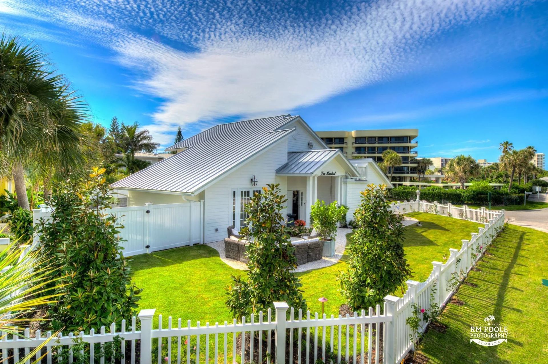 A white coastal-style house with a grey metal roof and white picket fence in a sunny, landscaped neighborhood.