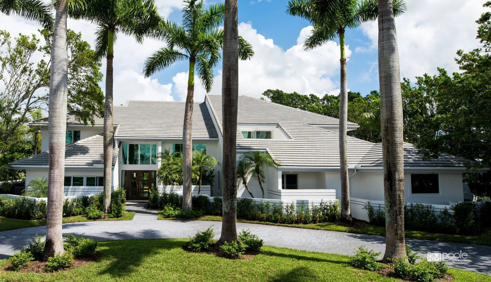 A large, multi-story white house with a modern gray roof, surrounded by tall palm trees and lush green landscaping.