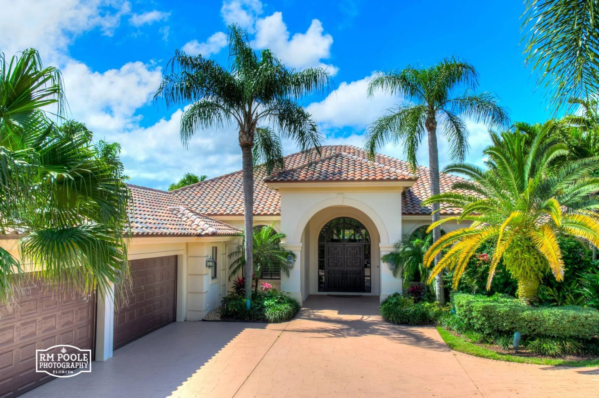 Single-story stucco home with a terracotta tile roof, arched entryway, and lush palm trees against a bright blue sky.