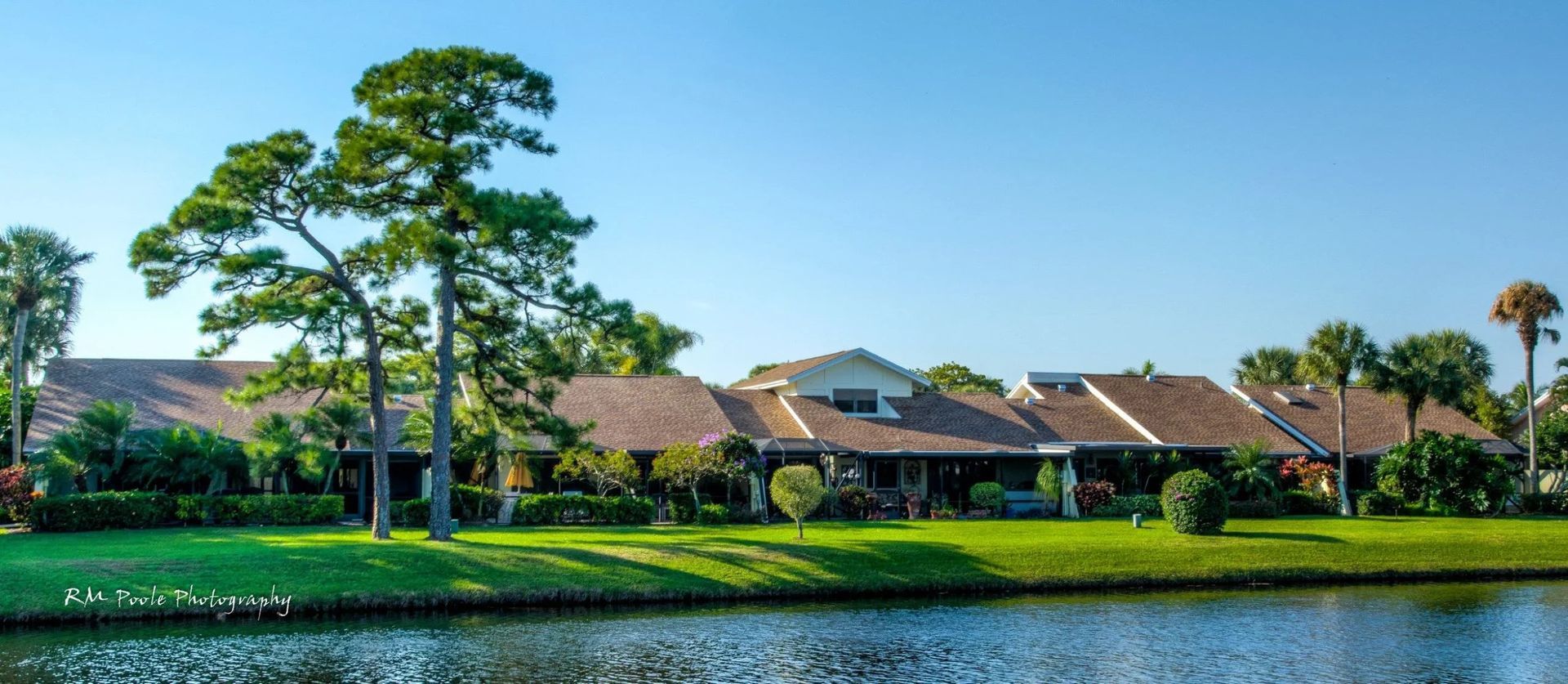 A row of houses with brown roofs sits behind a grassy bank next to a calm lake under a clear blue sky.