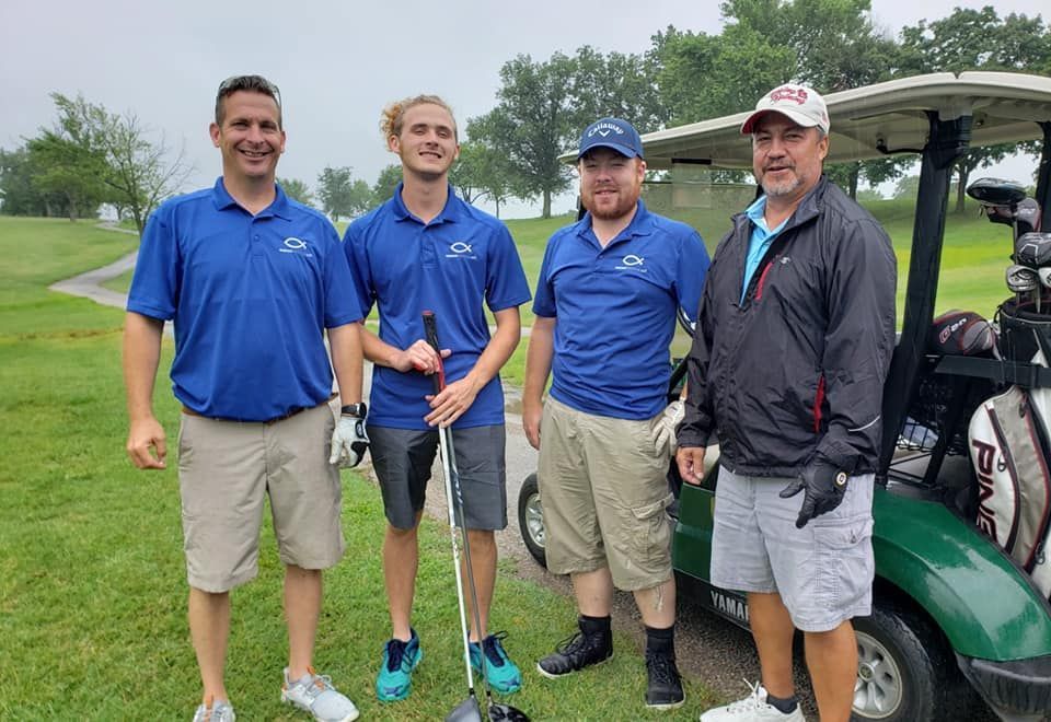 Four people stand in blue polo shirts and shorts on a golf course, posing for a photo beside a golf cart.