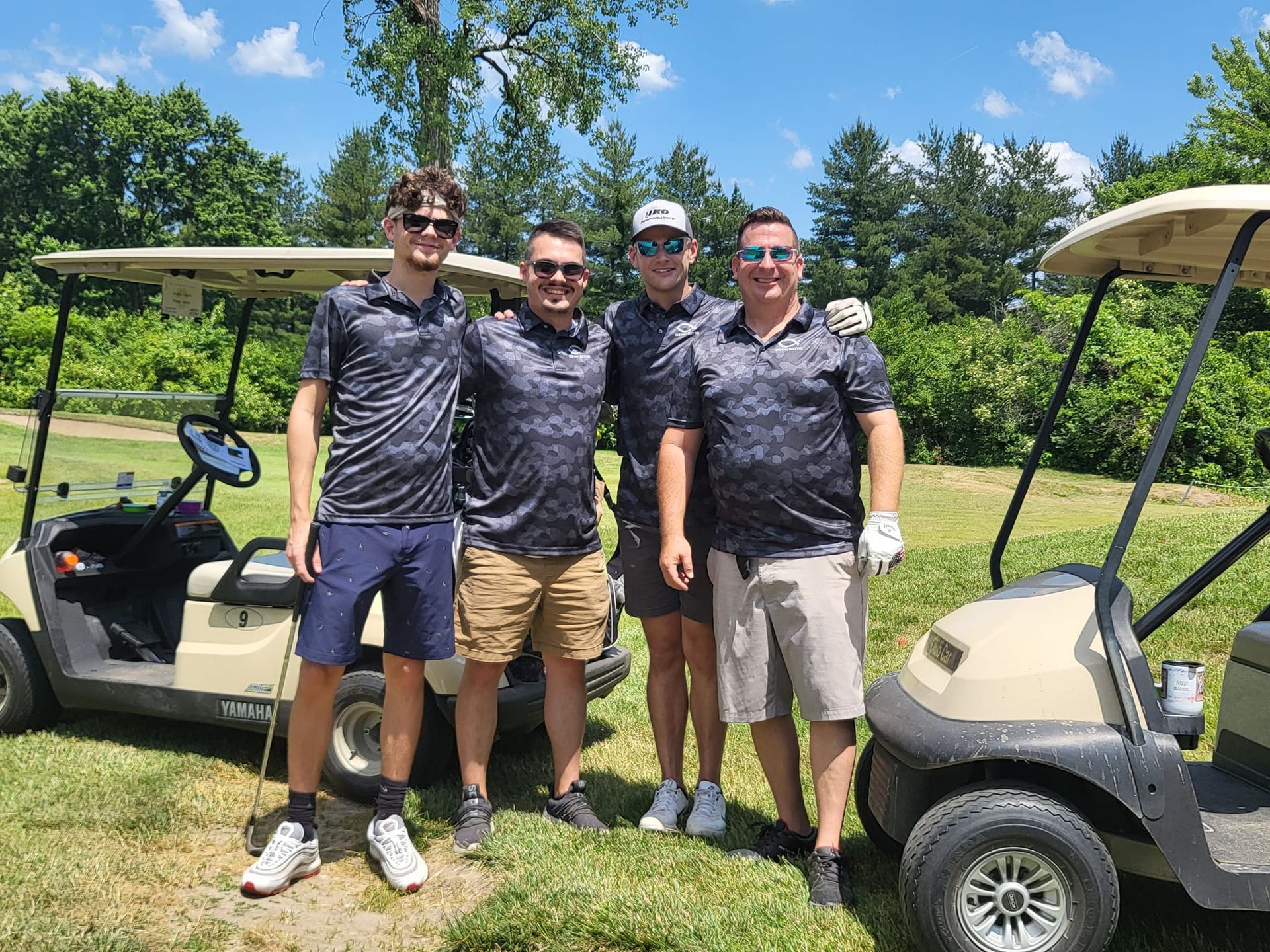 Four people wearing matching camouflage golf shirts stand outdoors between two golf carts on a sunny day.