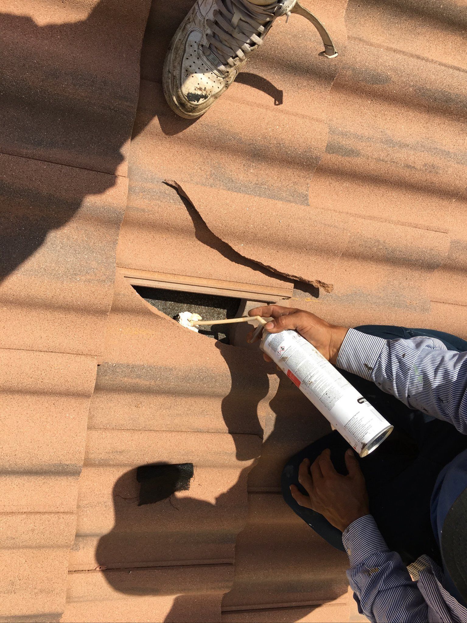 A person applies spray foam into a gap in terracotta-colored roof tiles.