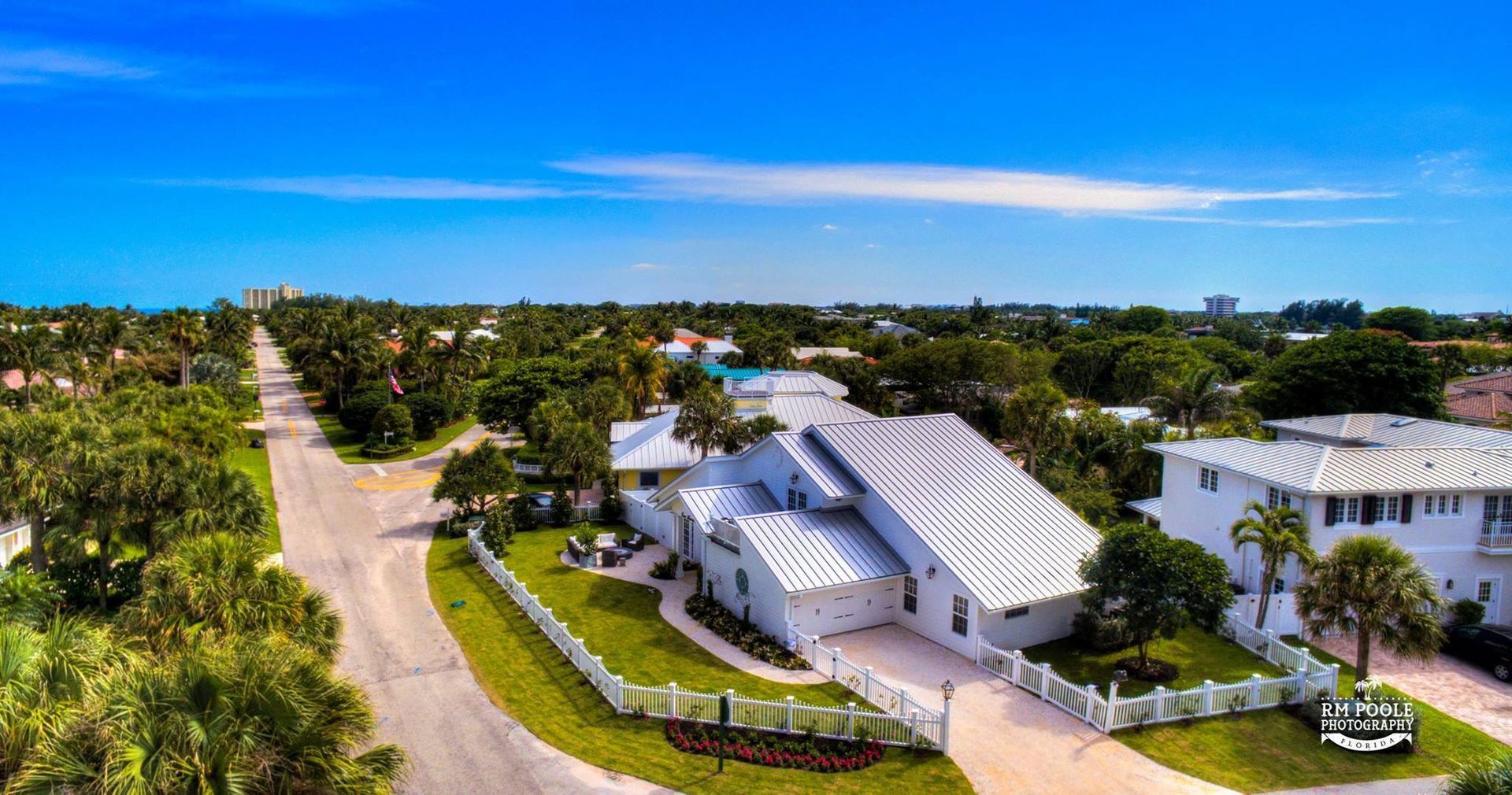Aerial view of a white, multi-roofed suburban home on a sunny day with lush green landscaping and a nearby street.